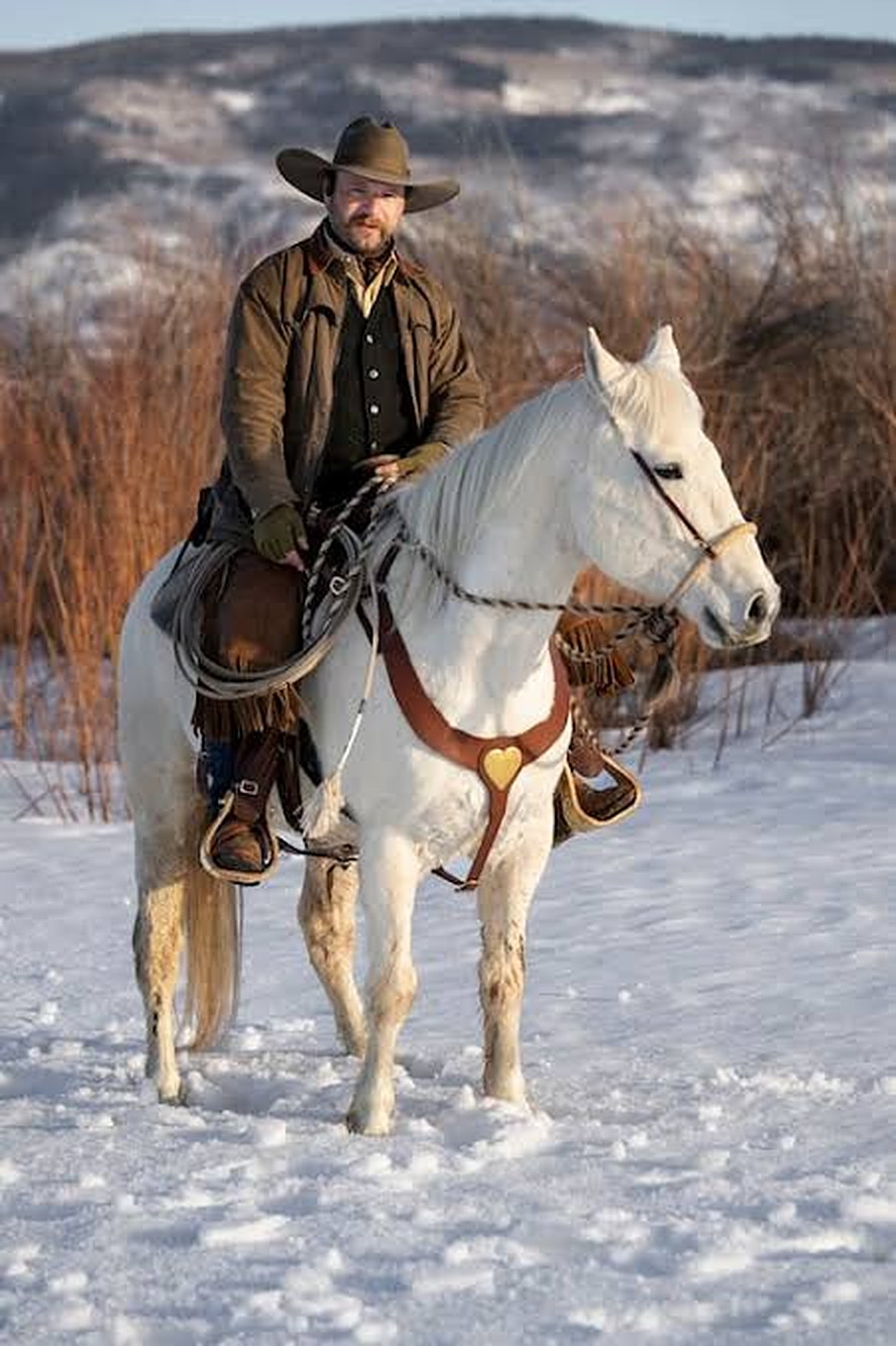 Cowboy in a brown hat and duster on a white horse in the snow